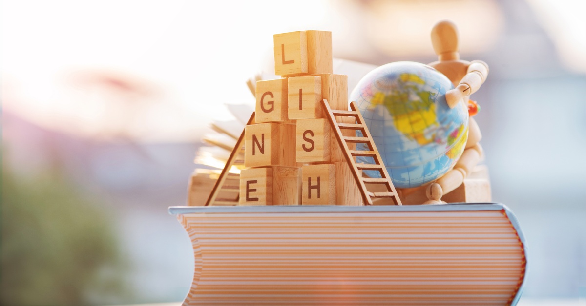 Wooden lettered blocks spelling the word "English" and a small wooden figure holding a globe, on top of a thick book.