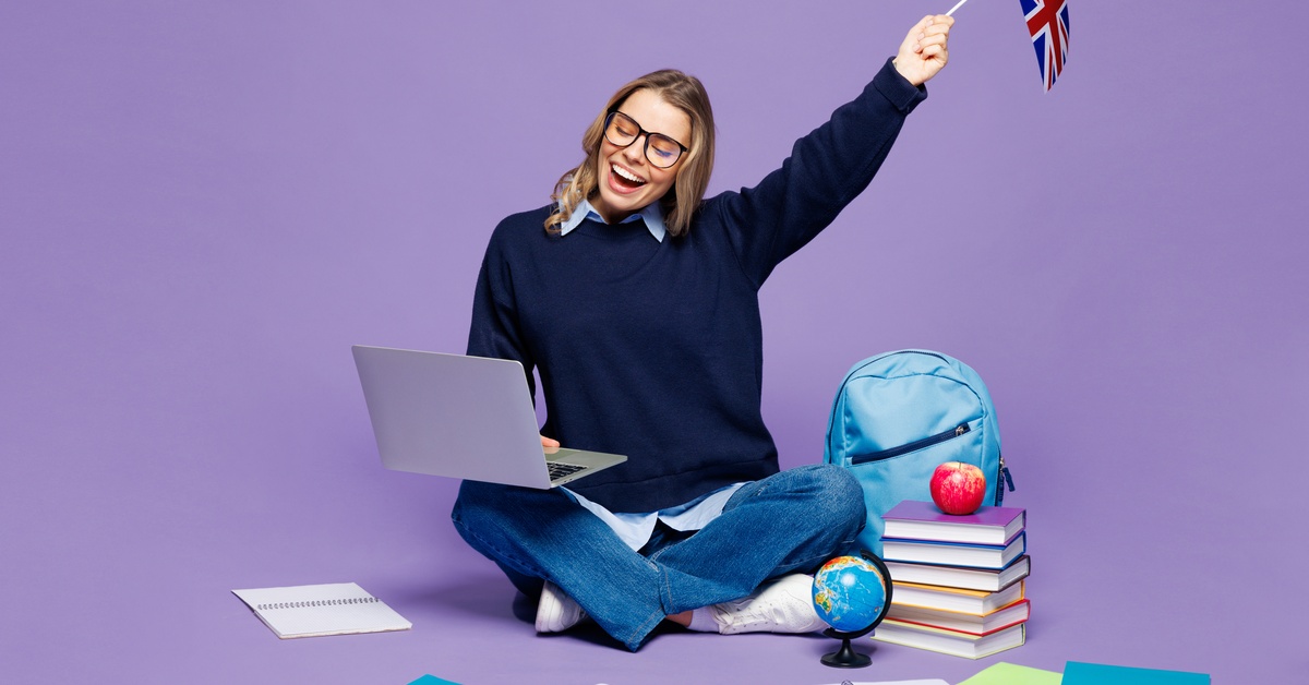 A woman wearing glasses sits next to papers and books with a laptop on her knee. She raises a small flag of the United Kingdom.