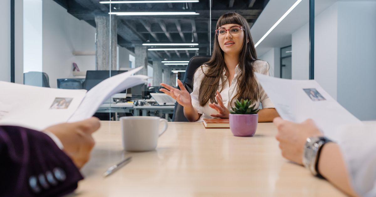 A woman wearing glasses sits at a table in an office. She sits opposite two other people with documents in their hands.