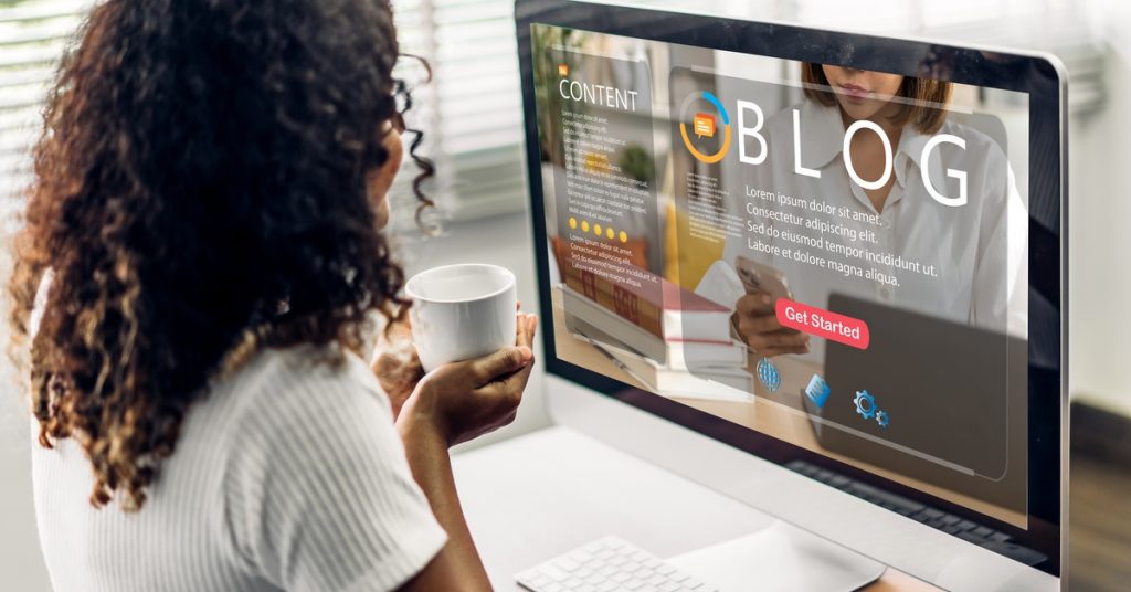 A woman holds a white cup as she sits at a desk in front of a computer screen that displays a blog page template.
