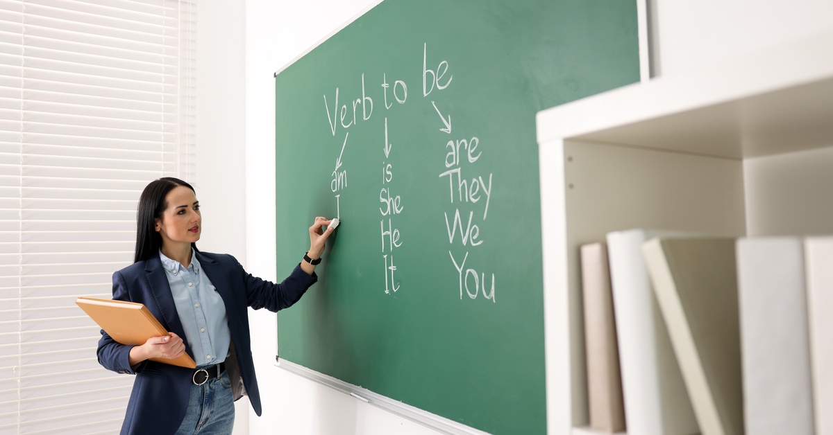 A woman holding chalk in her left hand and marking a chalkboard with words and arrows explaining the verb "to be" on it.