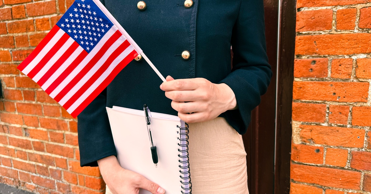 A person standing against the surface of a brick wall is holding a notebook in one hand and the US flag in the other.