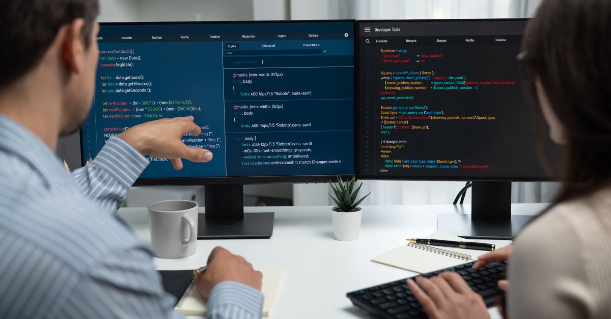 Two people sitting at a desk in front of keyboards and computer screens that display several lines of code.