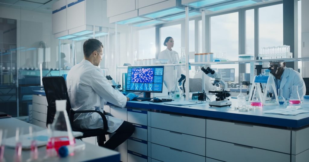 Two people wearing white coats in a laboratory. One person stands, while the other person sits and uses a computer.