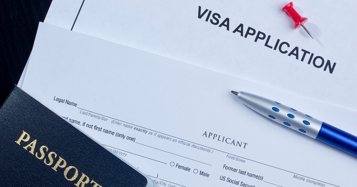 A blue passport, blue ink pen, and red push pin sitting on the surface of two documents for visa application.