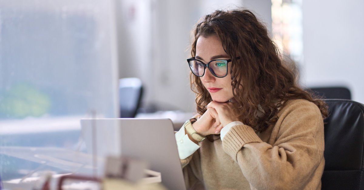 An individual wearing glasses and a light brown sweater sitting down and looking ahead at a laptop screen.