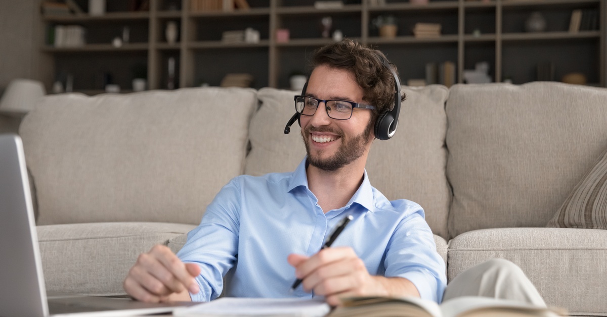 A smiling person wearing a microphone headset sits in front of a laptop with books on the table and a pen in his hand.