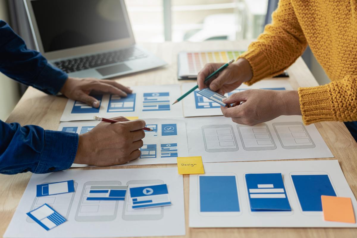 Two people stand across from each other and work together to develop a website, with concepts laid out on paper between them.