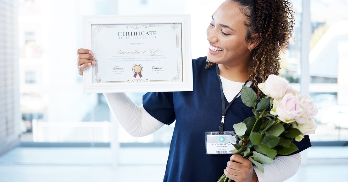 A woman wearing a blue and white healthcare uniform holds up a certificate in one hand and a bouquet in the other hand.