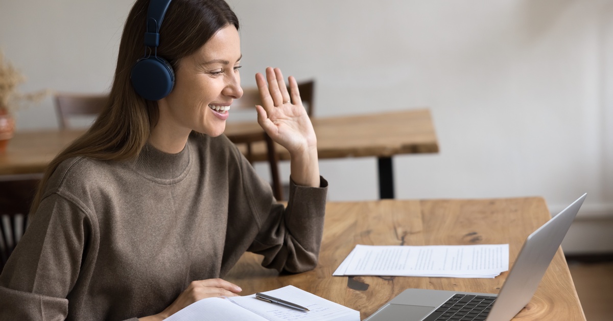 A smiling and happy young adult womanfemale in headset, waving hello to the meeting on her notebook tablet.