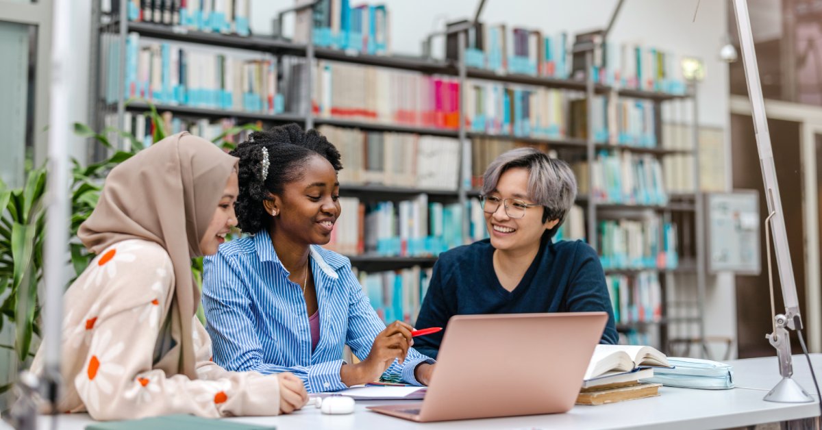 Multiethnic group of students sitting in a library and studying together and smiling at one another.
