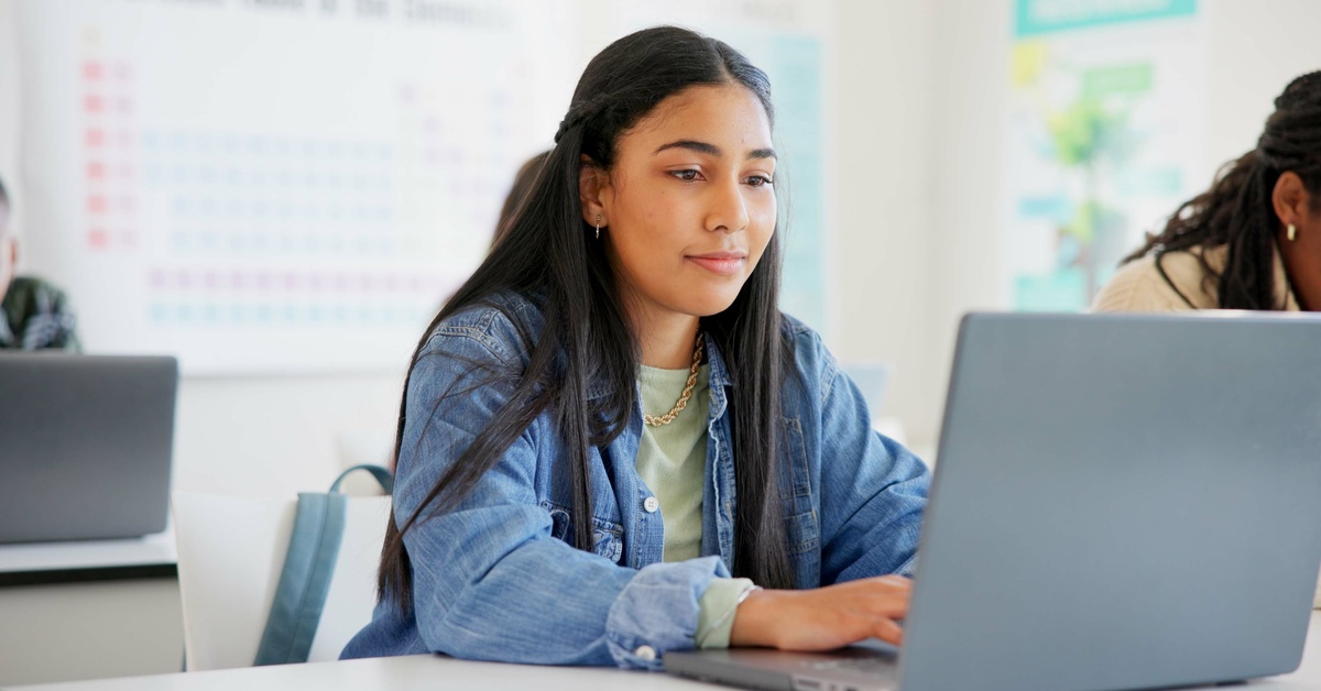 A woman is using a laptop in a classroom next to another student. She's concentrated on her own screen.