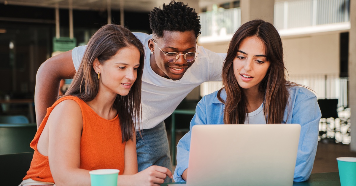 A group of three diverse happy students using a laptop for a university assignment in the college library.