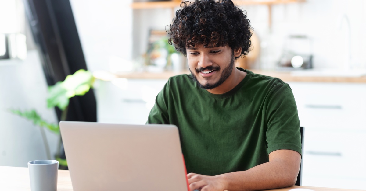 A happy young man smiles as he uses a laptop and an open notebook to work on schoolwork remotely from home.