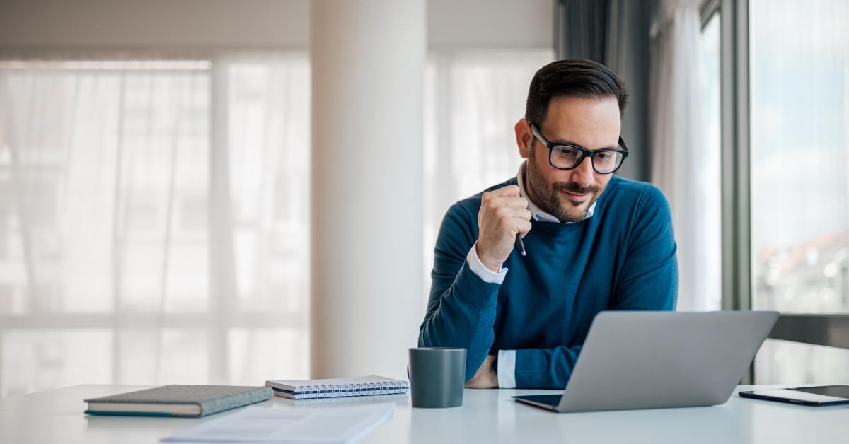 A serious businessman is working on a laptop at his office desk with coffee cups and notebooks next to him.