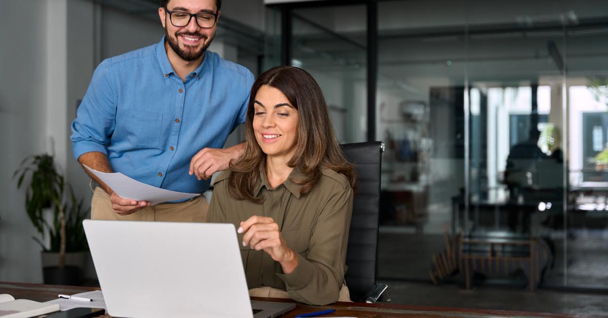Two happy professional business people are sharing notes and using a laptop while working together in the office.