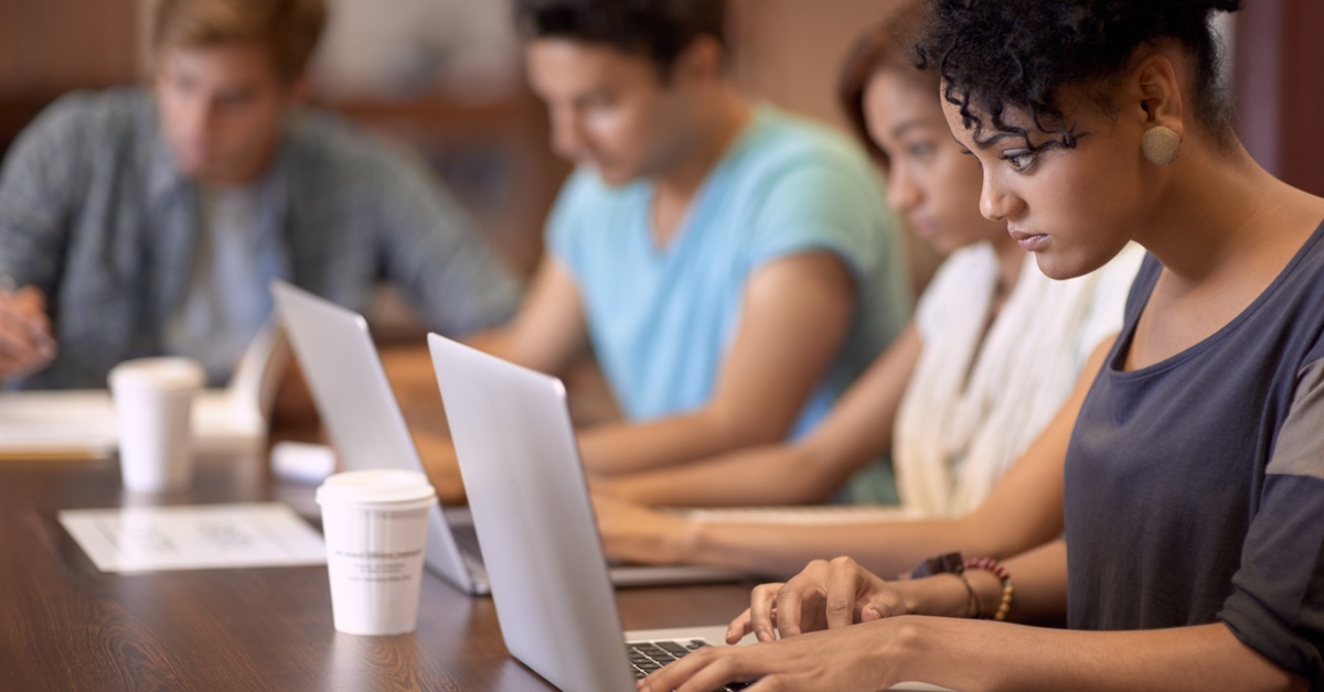 A close-up of a young woman and a group of students studying at a wooden desk in the library, using their laptops.