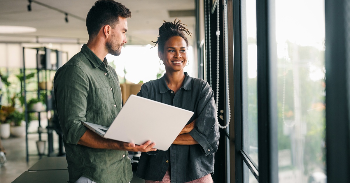 Two young professional software engineers are talking and gazing out the window while one holds a laptop.