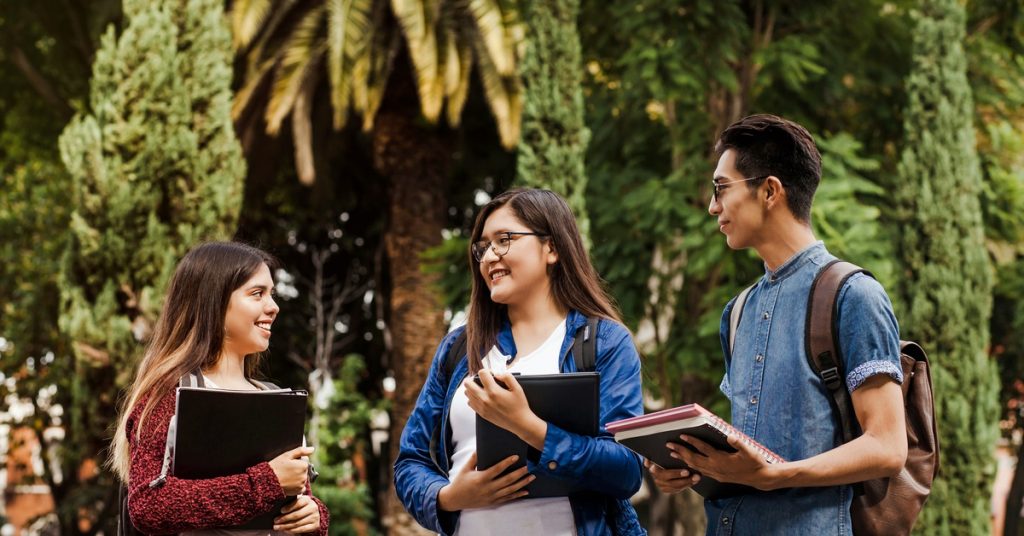 A group of three students is talking to each other on a college campus while holding notebooks and binders.