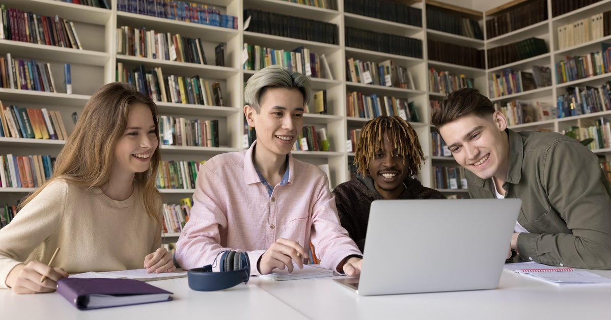 A group of diverse university students collaboratively studying in an empty library room and smiling at a laptop.