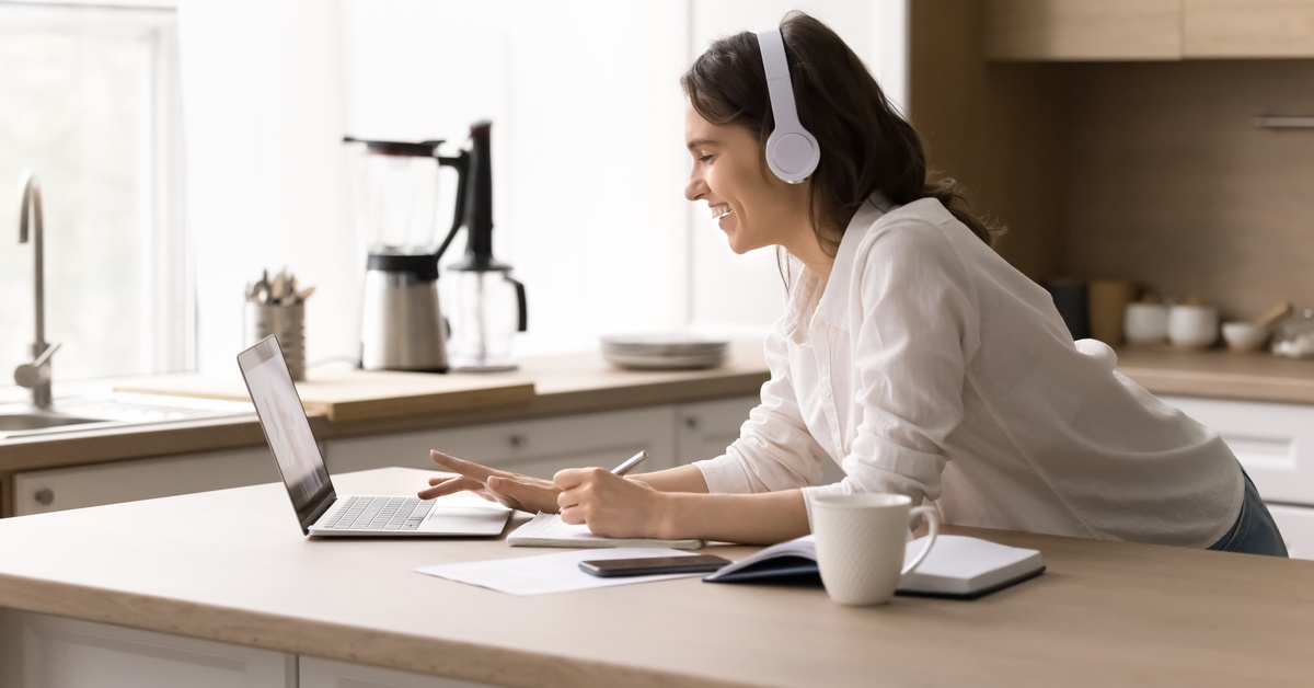 A close-up of a woman using her laptop and Bluetooth headphones in her kitchen. She's smiling at the computer.