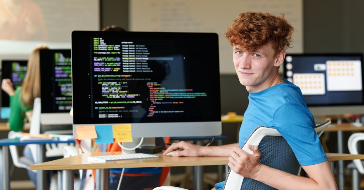 A male student with ginger hair is looking at the camera while using a computer at an IT secuirty lesson.
