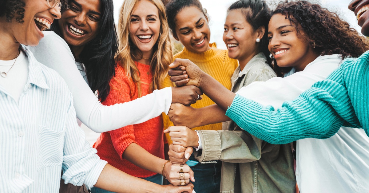 A multicultural group of women stacking hands together and laughing with each other while standing in a circle.