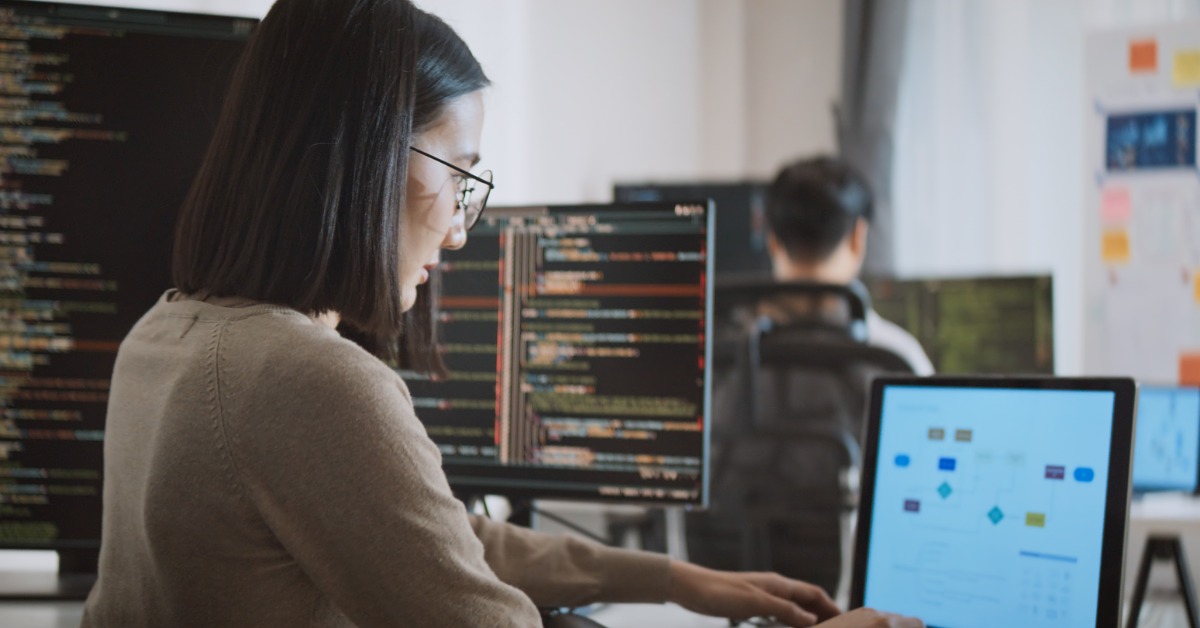 Woman wearing glasses works at laptop in office; code fills dual monitors behind, coworker blurred at desk.