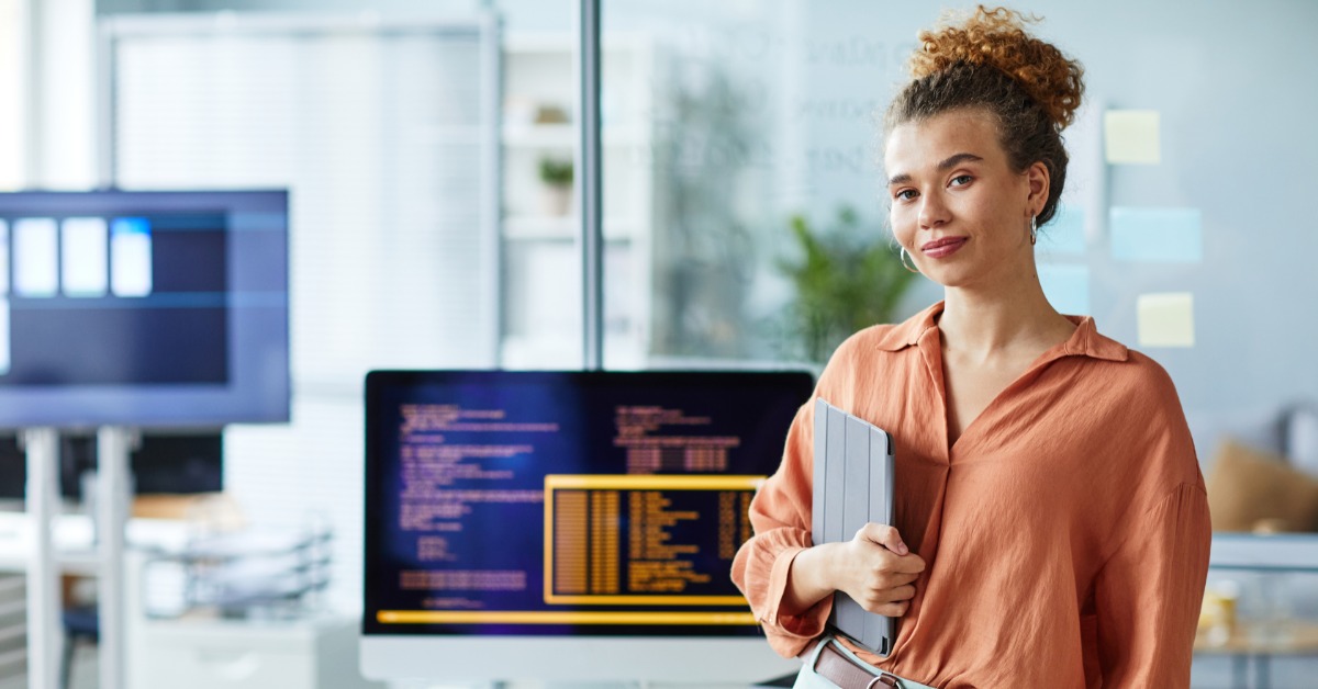 Developer with curly hair holds a laptop by her desk; monitor behind shows code with yellow highlighted panel.