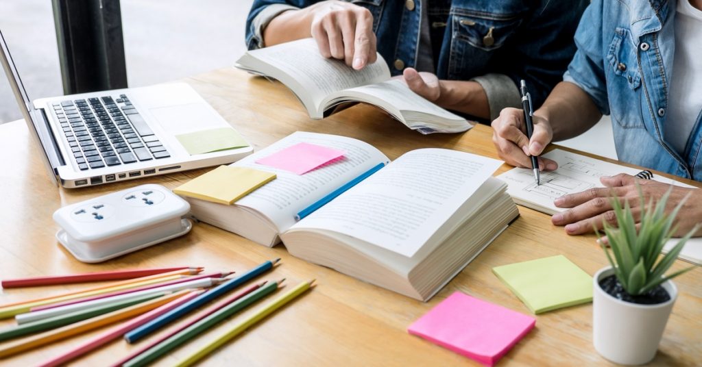 Two classmates are studying, reading, and doing homework at a wooden desk to help prepare for an exam.