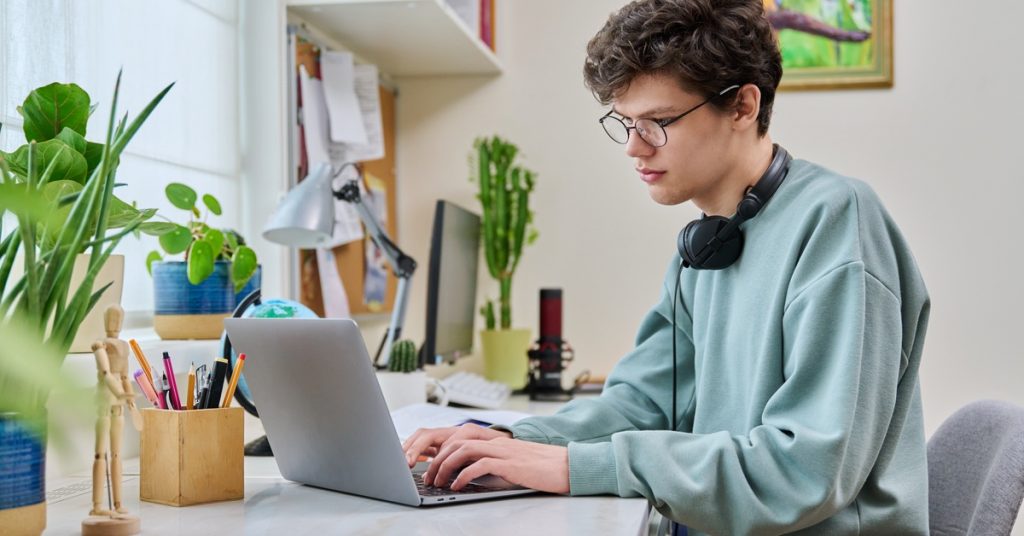 A young male college student is sitting at a desk at home using a laptop with headphones draped around his neck.