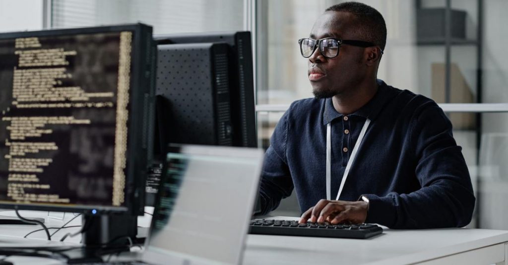 Man wearing glasses coding at desktop computer, lines of software code visible on monitor in modern office.