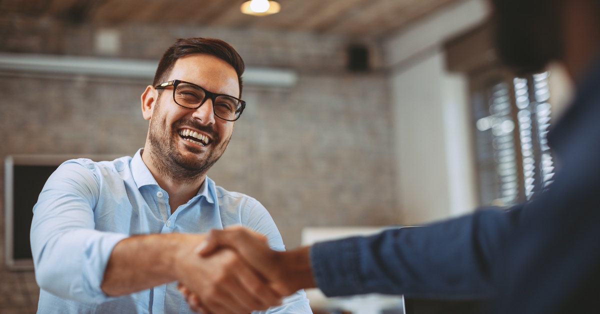 Smiling businessman wearing glasses shakes hands with a colleague across a table in a modern office meeting.