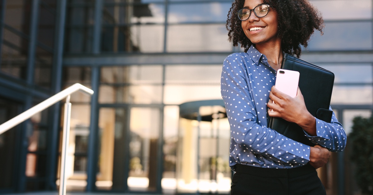 Businesswoman with curly hair and glasses stands outside an office building, smiling, holding a phone and laptop folder.
