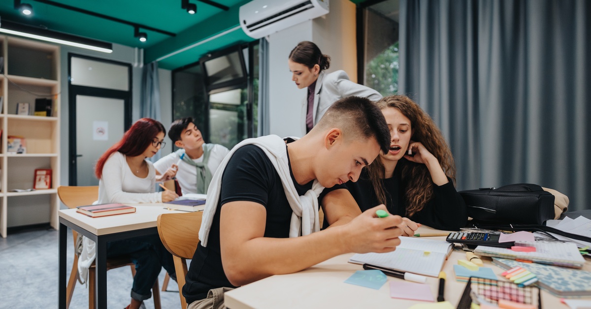 A group of students studying together at desks in a classroom while focusing on notes, homework, and school assignments.