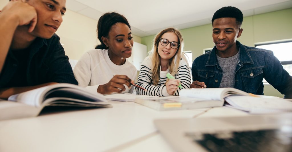 A group of diverse college students studying together at a table, discussing ideas and reviewing textbooks in a classroom.