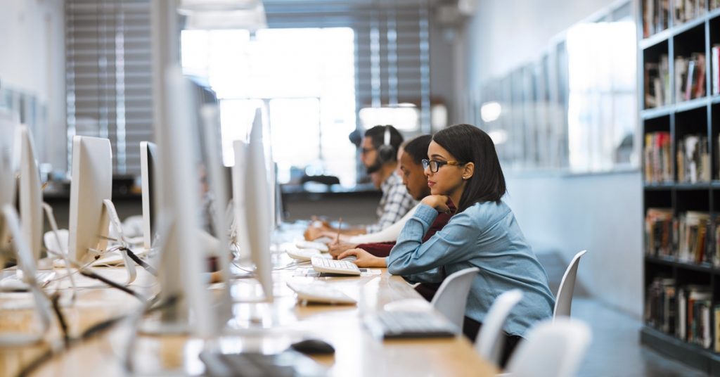 People working at computers in a study space, seated in a row with monitors and bookshelves in the background