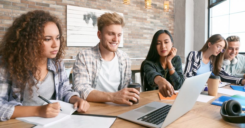 Young adults study together around laptop, writing notes and discussing while seated at wooden table indoors.