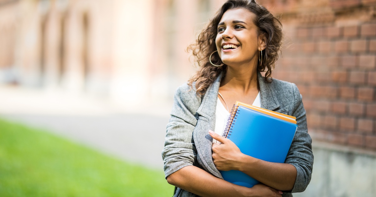 Smiling woman outdoors holds notebooks against chest, looking aside near brick wall and grassy area in soft daylight.