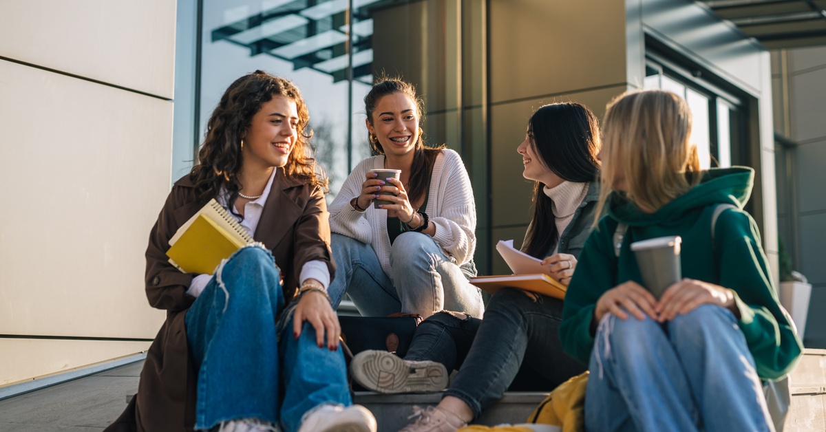 Four young women sit on steps outside building, chatting and holding notebooks and coffee in warm sunlight.