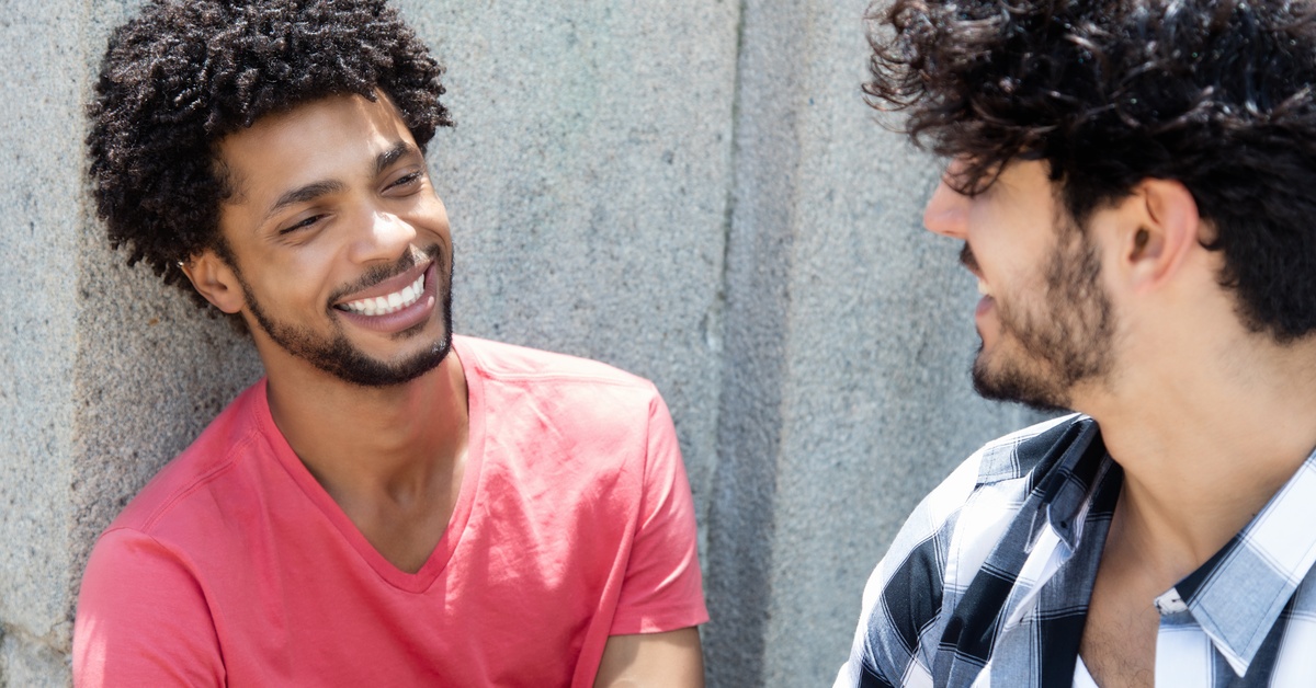 Two male friends chat and smile while seated outdoors by wall, casual clothing and relaxed conversation captured.