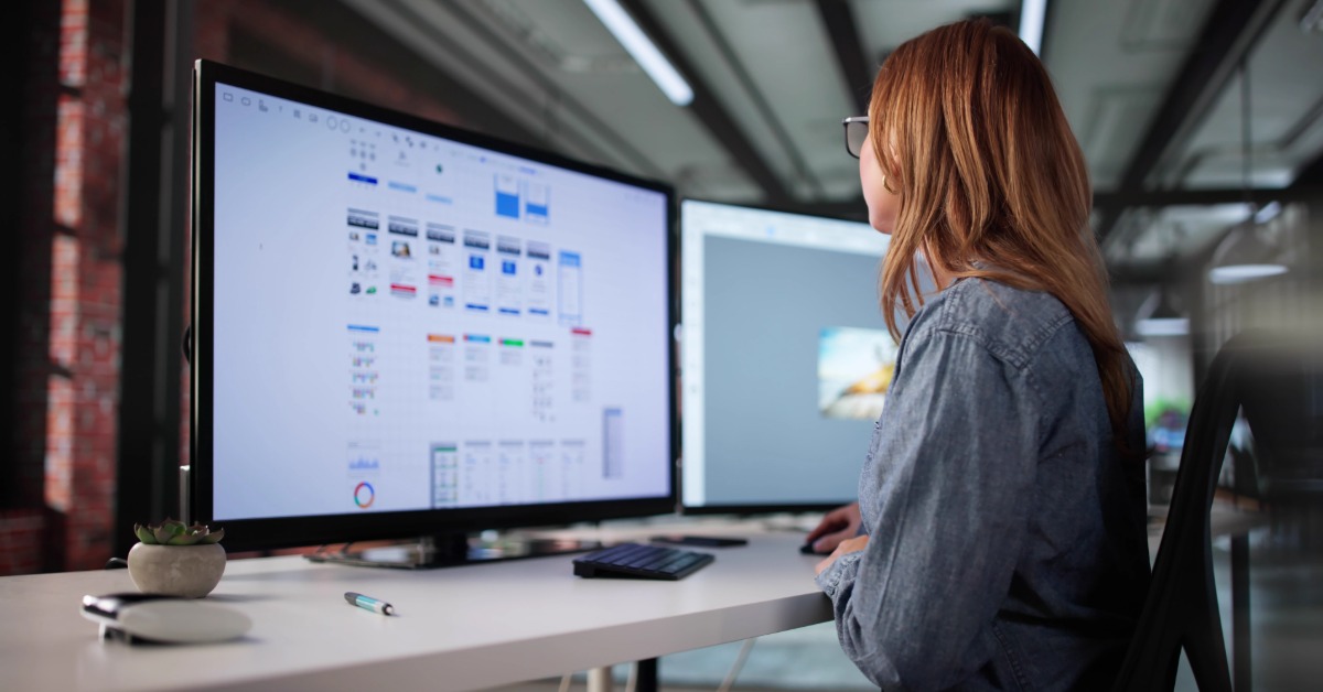 Designer works at dual monitors reviewing interface layouts and graphics, seated at desk in modern workspace.