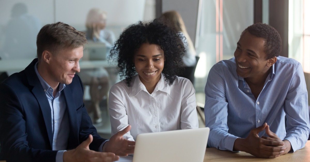 Three coworkers smile while reviewing laptop together at office table, discussing ideas in bright meeting space.