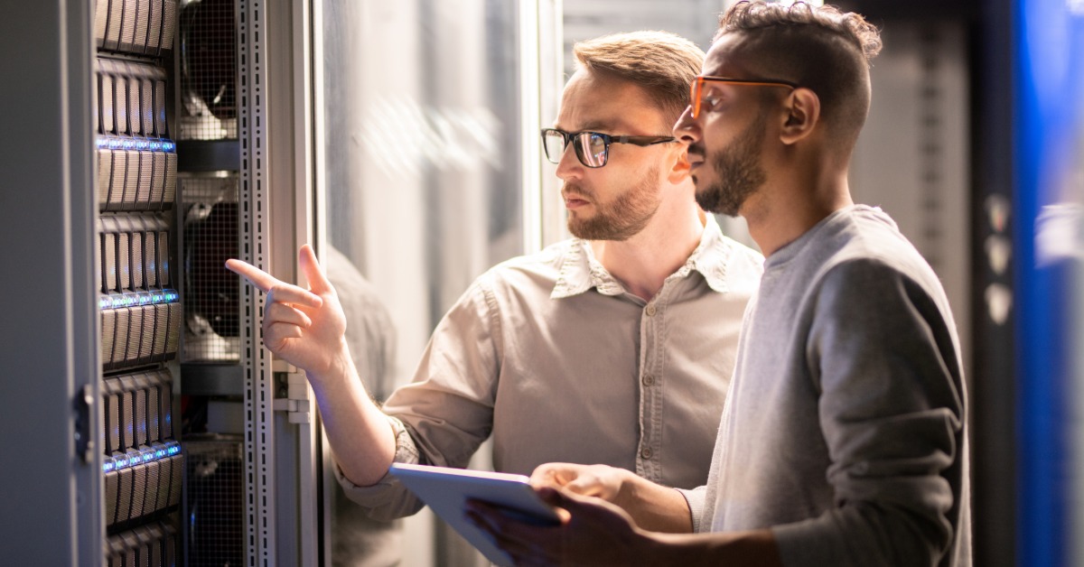 IT professionals working together in server room, reviewing hardware while using tablet beside network equipment racks.