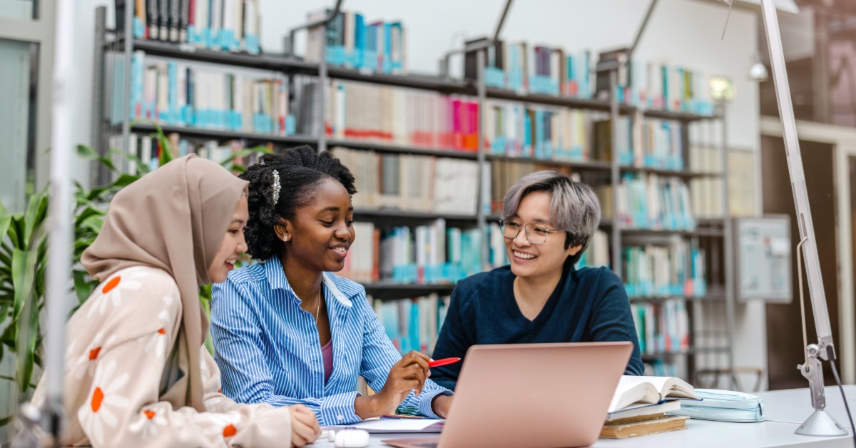 Three classmates collaborate at a library table with a pink laptop, notebooks, open books, and shelves in the background.