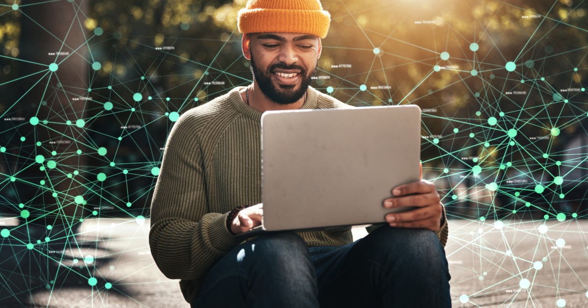 Man in an orange knit hat uses a laptop outdoors, framed by glowing network lines and dots in sunlight.