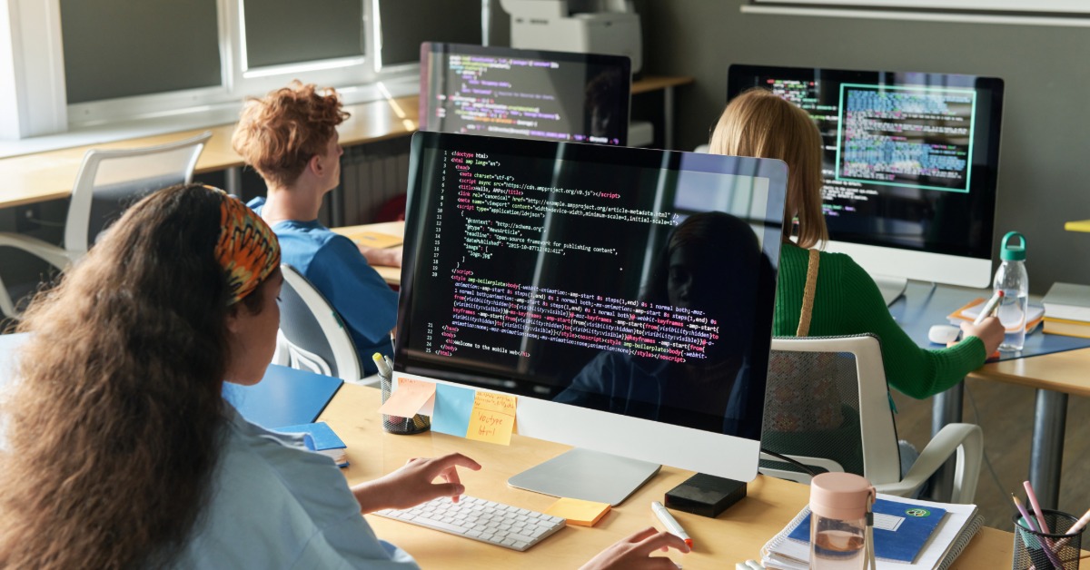 A classroom of students studies programming on large monitors displaying code, with supplies arranged on desks.