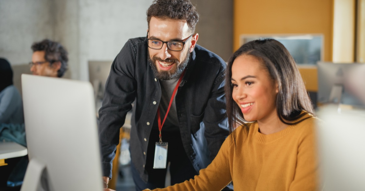 Two coworkers smile while looking at a desktop computer in a bright office, with another person working behind them.