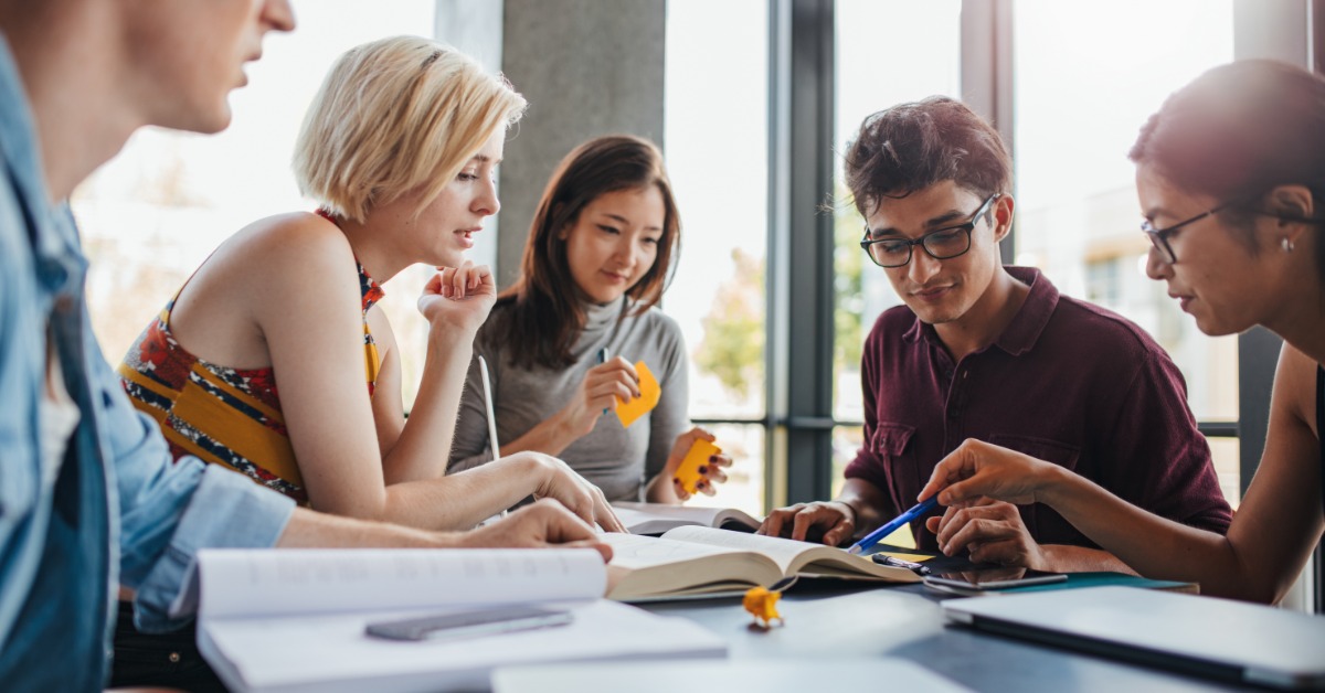 Group of students studies together with textbooks, notebooks, and sticky notes beside large sunlit windows.