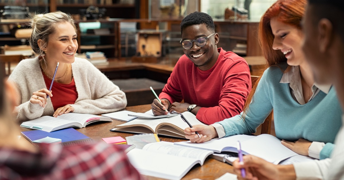 Students smile and take notes around a library table with open books, pencils, and study materials spread out.